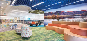 Interior view of a library children's area with curved tiered seating, photo mural, playful acoustic ceiling, and interactive Jeep play area