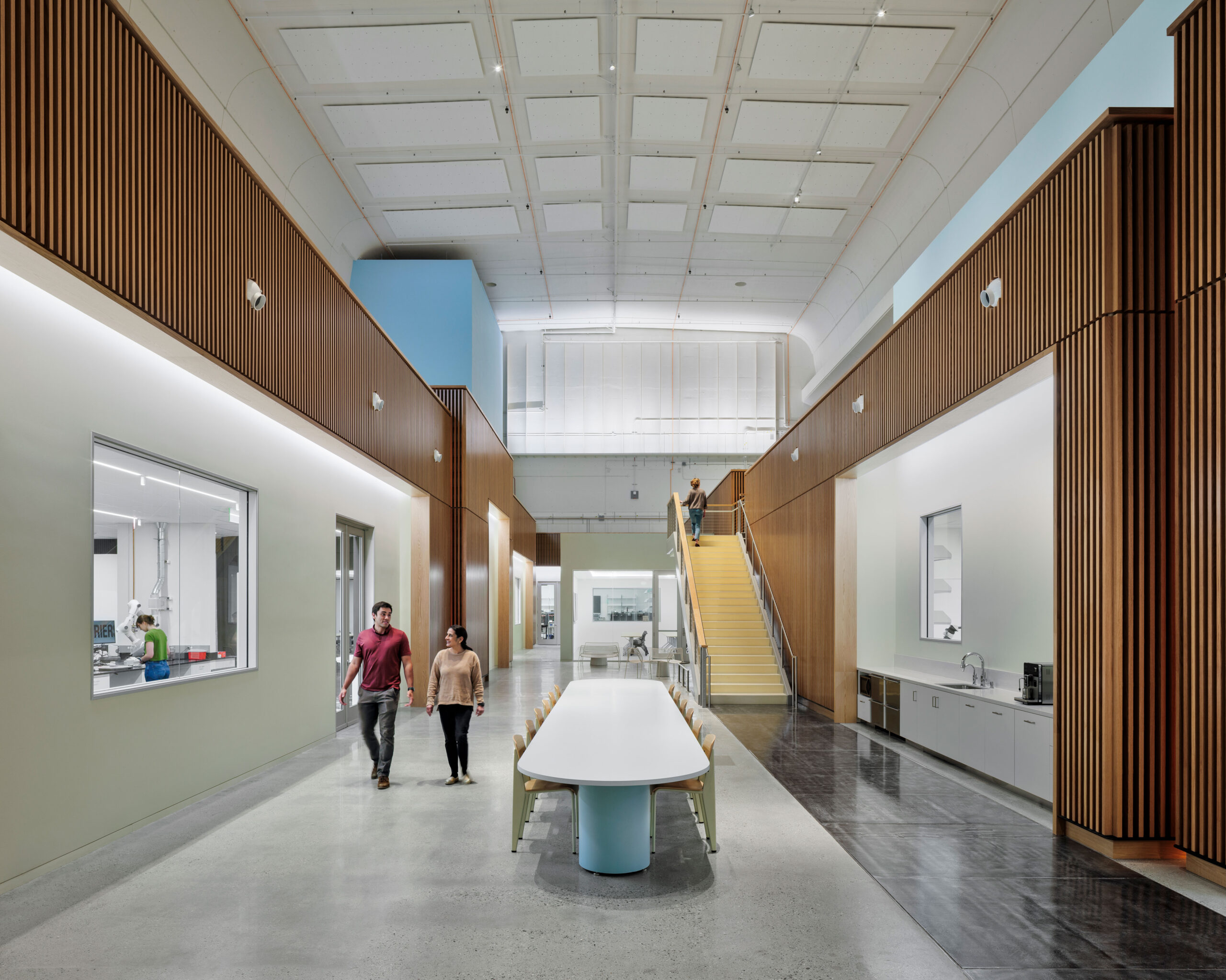 Double-height corridor in the Stanford University DEEP Lab, lined with wood-slat paneling and a glass-walled laboratory, connecting a shared collaboration table and kitchenette to a mezzanine stair above. A CAS Architects, an FPBA Co. and Jensen Architects education project.