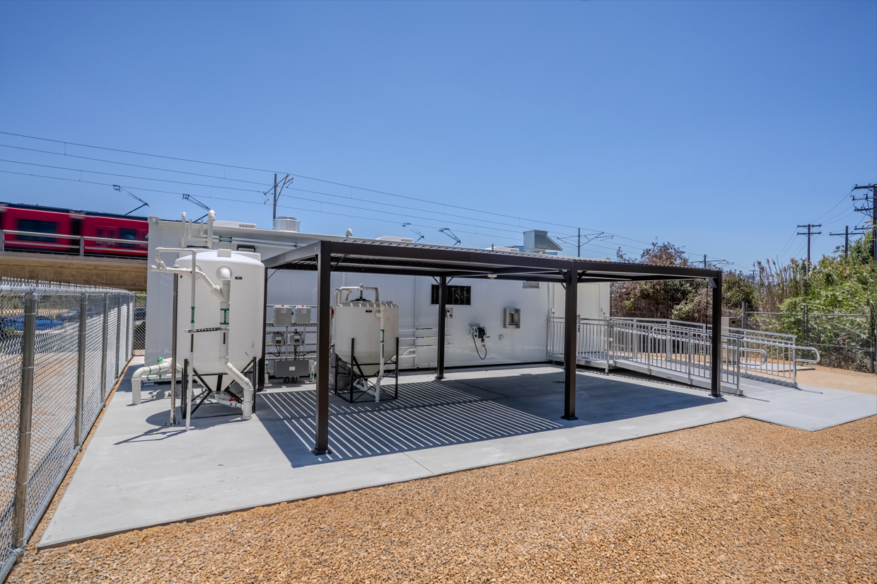 A trolley passes behind SDSU's One Water Laboratory, a modular field lab with a covered outdoor work area and accessible ramp. The lab is an FPBA higher education project for San Diego State University in Mission Valley.